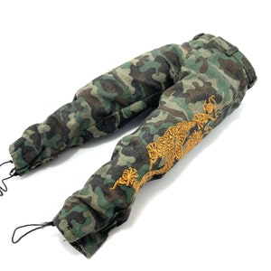 May include: Camouflage trousers with a gold embroidered design on the right leg. The trousers have a drawstring at the bottom of each leg.