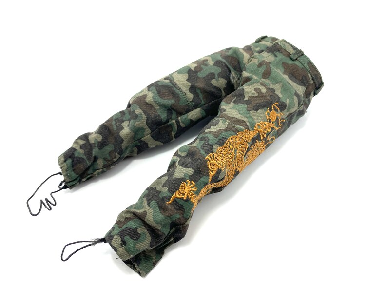 May include: Camouflage trousers with a gold embroidered design on the right leg. The trousers have a drawstring at the bottom of each leg.