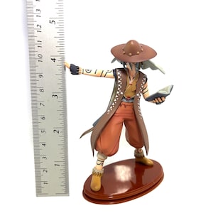 May include: A detailed figurine of a character in a brown hat, vest, and pants, holding a book and gesturing with one arm. The figure is approximately 4 inches tall and stands on a brown oval base. The figurine has intricate details and is set against a white background.