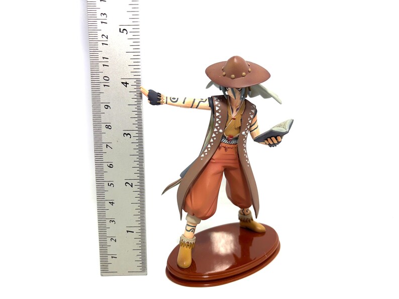 May include: A detailed figurine of a character in a brown hat, vest, and pants, holding a book and gesturing with one arm. The figure is approximately 4 inches tall and stands on a brown oval base. The figurine has intricate details and is set against a white background.