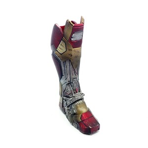 Pu&ograve; includere: Un pezzo di armatura per la gamba di Iron Man in metallo rosso, oro e argento. L'armatura ha un aspetto usurato con linee di pannelli e dettagli meccanici visibili, che suggeriscono un'estetica usurata dalla battaglia.