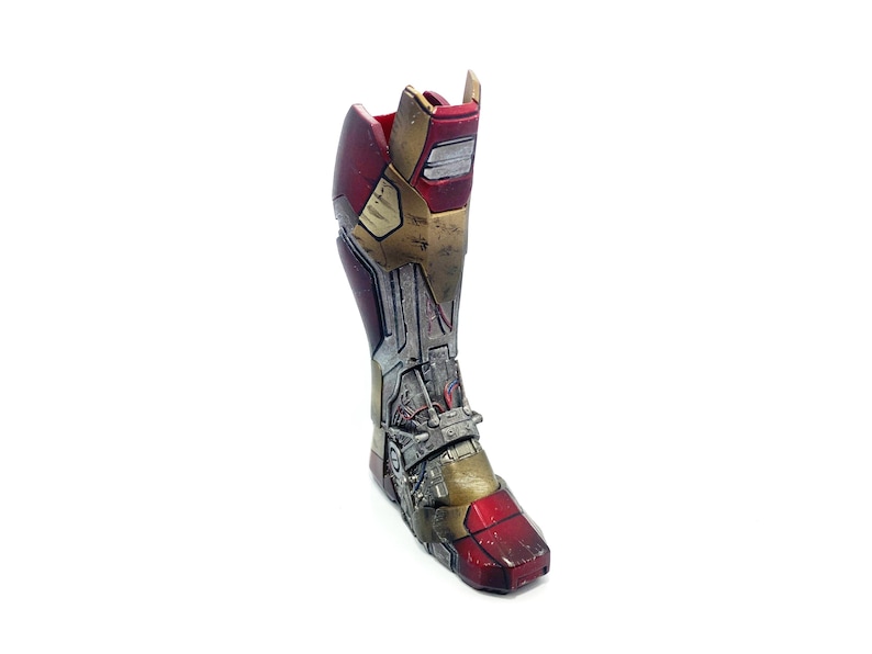 Pu&ograve; includere: Un pezzo di armatura per la gamba di Iron Man in metallo rosso, oro e argento. L'armatura ha un aspetto usurato con linee di pannelli e dettagli meccanici visibili, che suggeriscono un'estetica usurata dalla battaglia.
