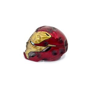 May include: A red and gold Iron Man helmet replica. The helmet is a detailed miniature, featuring the iconic design with a metallic red finish and gold accents. The helmet is a collectible item.