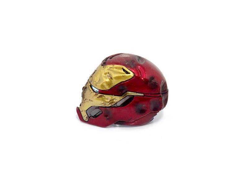 May include: A red and gold Iron Man helmet replica. The helmet is a detailed miniature, featuring the iconic design with a metallic red finish and gold accents. The helmet is a collectible item.