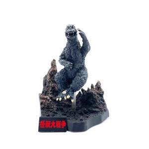 May include: A gray Godzilla figurine sitting on a black base with a rocky landscape. The base has a black plaque with the Japanese text "怪獣大戦争" in red.