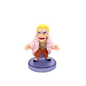 May include: A figurine of a character with yellow hair, sunglasses, and a pink feathery coat. The figure is wearing an orange outfit and black shoes, standing on a purple base with the text "Doflamingo".