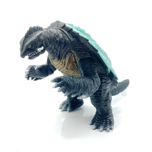 Pode incluir: Figura detalhada de um monstro semelhante a uma tartaruga, predominantemente cinza escuro com uma carapaça verde-azulado claro. A criatura tem dentes afiados, olhos verdes e um corpo texturizado. Está em pé sobre quatro patas, com os braços levantados.