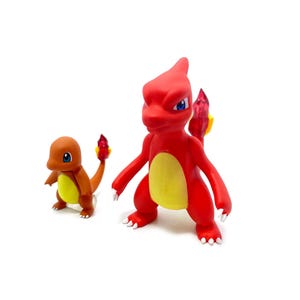 Puede incluir: Dos figuras de Pokémon rojas y naranjas. La figura más grande es un Charmeleon con la barriga amarilla y una cola roja con una llama. La figura más pequeña es un Charmander con la barriga amarilla y una cola naranja con una llama.