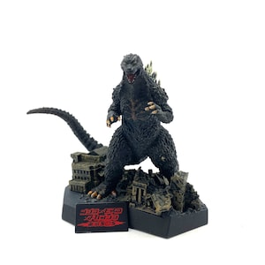 May include: A black Godzilla figure standing on a destroyed city base. The figure is posed with its mouth open and its arms raised. The base is made of gray and brown plastic and features a small sign with the text "ゴジラ キング オブ モンスターズ" in Japanese.