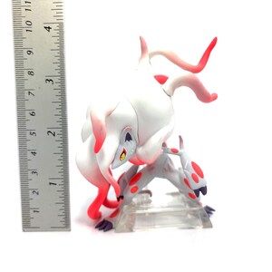 Puede incluir: Figura blanca y roja de una criatura m&iacute;tica, de unos 10 cm de altura, en una pose din&aacute;mica. La figura tiene un cuerpo blanco con detalles rojos y est&aacute; sobre una base de pl&aacute;stico transparente. Una regla est&aacute; al lado.