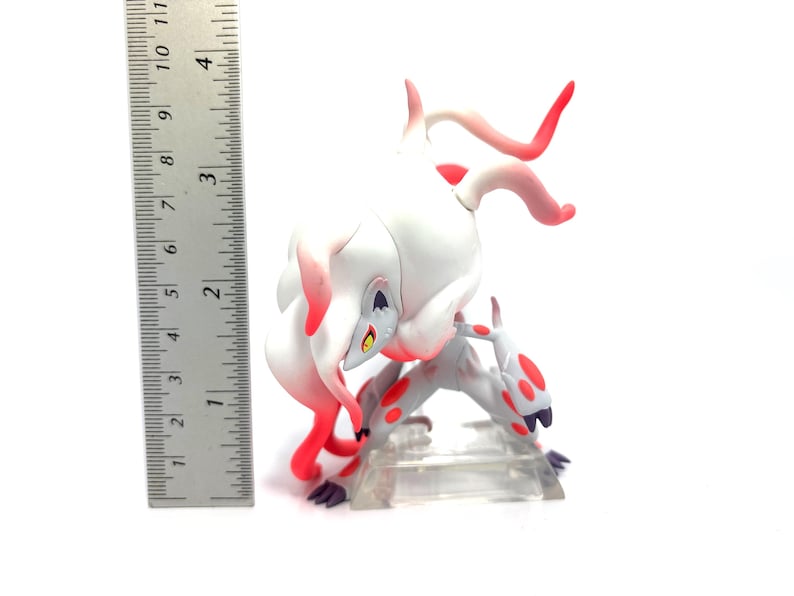 Puede incluir: Figura blanca y roja de una criatura m&iacute;tica, de unos 10 cm de altura, en una pose din&aacute;mica. La figura tiene un cuerpo blanco con detalles rojos y est&aacute; sobre una base de pl&aacute;stico transparente. Una regla est&aacute; al lado.