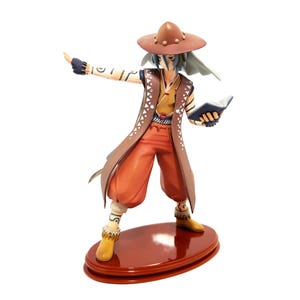 May include: A figurine of a character in a brown hat, vest, and pants, holding a book and pointing. The figure has blue hair and is standing on a brown oval base. The outfit includes a long brown coat and yellow boots.