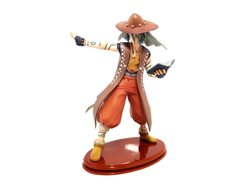 May include: A figurine of a character in a brown hat, vest, and pants, holding a book and pointing. The figure has blue hair and is standing on a brown oval base. The outfit includes a long brown coat and yellow boots.