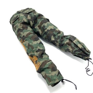 May include: Camouflage trousers with a drawstring at the bottom of the legs. The trousers have a gold embroidered design on the left leg.