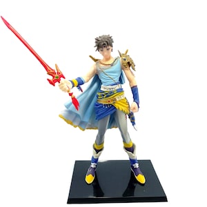 Könnte beinhalten: Eine blaue und gelbe Figur eines männlichen Charakters, der ein rotes Schwert hält. Der Charakter trägt eine blaue und gelbe Tunika und einen blauen Umhang. Die Figur steht auf einem schwarzen Sockel.