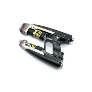 May include: A black and silver toy gun with a carbon fiber pattern. The gun has a gold-colored barrel and a silver trigger.