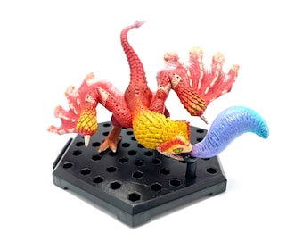 May include: A detailed figurine of a red, yellow, and blue mythical creature with a long neck and tail, perched on a black hexagonal base. The creature has detailed wings and a long, curved tail. The figurine is set against a white background.