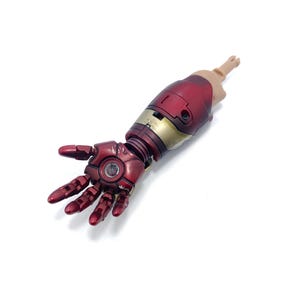 K&ouml;nnte beinhalten: Detaillierte Nachbildung eines Iron Man-Arms mit metallisch roter und goldener Farbgebung. Der Arm umfasst eine Hand mit beweglichen Fingern und einen Unterarm mit komplizierten mechanischen Details. Das Modell ist vor wei&szlig;em Hintergrund.