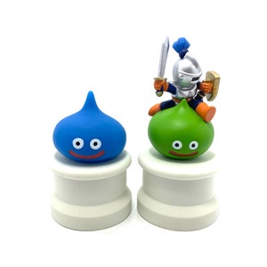 Puede incluir: Dos figuras de slime azul y verde con caras sonrientes sobre pedestales blancos. Una pequeña figura de caballero con una espada y un escudo está montada en el slime verde.