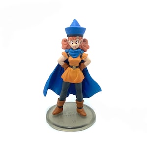 Puede incluir: Una figura de un personaje femenino de color azul y naranja que lleva un sombrero y una capa azules. La figura está de pie sobre una base redonda de plata.