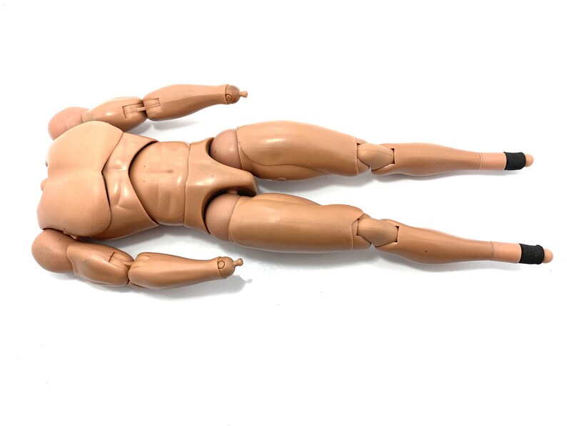 May include: A tan-colored articulated action figure body, lying horizontally. The figure has a detailed, realistic design with visible joints at the elbows, knees, and other points. The feet have black bands. The figure is designed for customization.