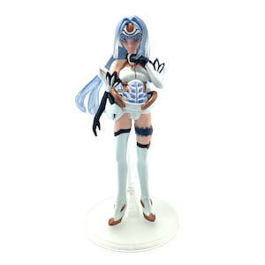 May include: A figurine of a female anime character with long, light blue hair and a futuristic outfit. The figure is predominantly white with accents of black, gold, and blue. It stands on a clear, circular base.