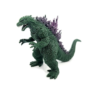 K&ouml;nnte beinhalten: Eine gr&uuml;ne Godzilla-Figur mit lila R&uuml;ckenflossen. Die Figur steht auf einer wei&szlig;en Oberfl&auml;che, mit offenem Maul und ausgestreckten Krallen. Die Figur hat ein strukturiertes, schuppiges Aussehen.