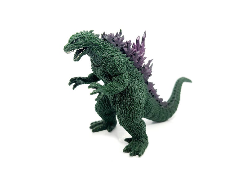 K&ouml;nnte beinhalten: Eine gr&uuml;ne Godzilla-Figur mit lila R&uuml;ckenflossen. Die Figur steht auf einer wei&szlig;en Oberfl&auml;che, mit offenem Maul und ausgestreckten Krallen. Die Figur hat ein strukturiertes, schuppiges Aussehen.