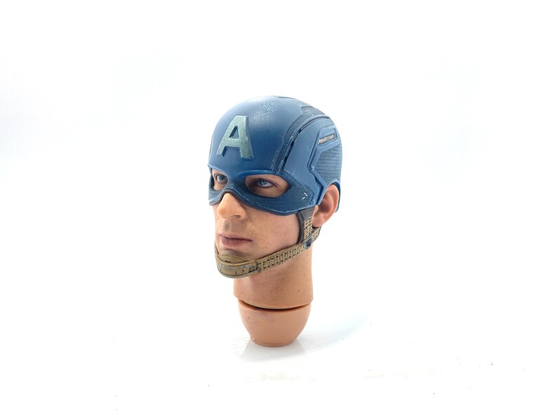 Op de afbeelding: Een gedetailleerde hoofdsnede van Captain America, met een blauwe helm met de letter "A" en een masker. De figuur heeft realistische huidtinten en een gebruinde nek. Een verzamelobject.