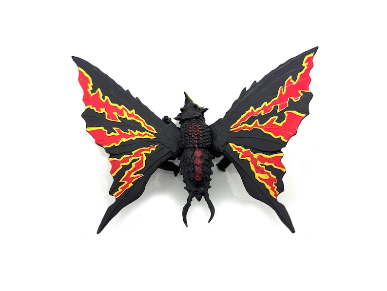 May include: A black creature figurine with large, wing-like appendages featuring vibrant red and yellow flame-like patterns. The central body has a textured, dark surface with a red stripe down the back and a small, pointed horn on the head.