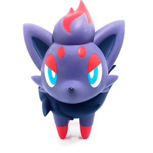 May include: A purple figurine of a Zorua character with red and blue accents. The figure has large ears, red eyes, and red feet. The top of the head has red flame-like details. The character is standing on a white surface.