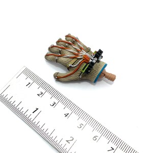 Puede incluir: Un guante beige y marr&oacute;n con cables naranja y blanco y un dispositivo negro y verde unido a la mu&ntilde;eca. El guante mide aproximadamente 7,5 cm de largo y est&aacute; sobre una superficie blanca junto a una regla.