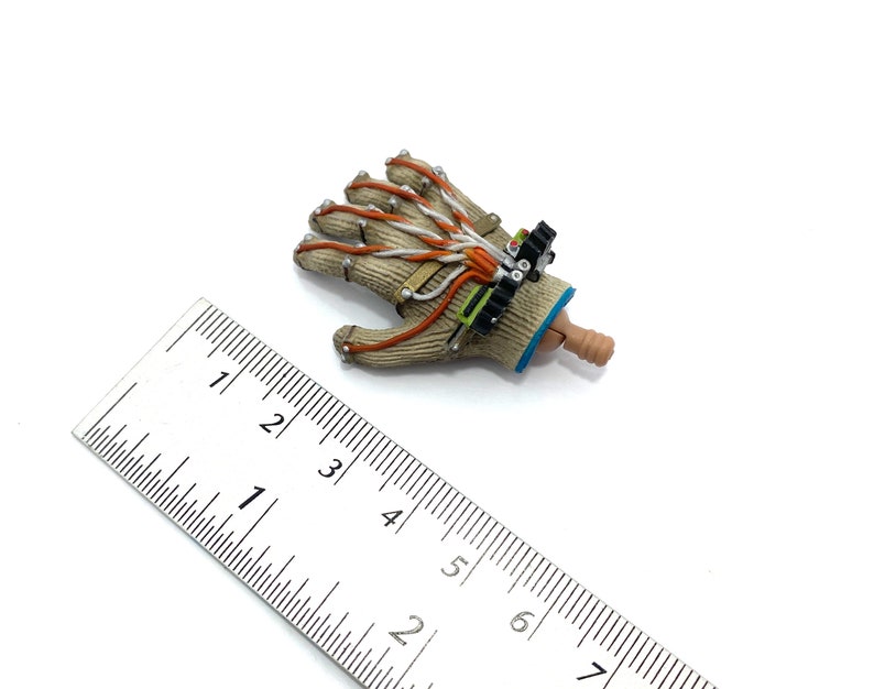 Puede incluir: Un guante beige y marr&oacute;n con cables naranja y blanco y un dispositivo negro y verde unido a la mu&ntilde;eca. El guante mide aproximadamente 7,5 cm de largo y est&aacute; sobre una superficie blanca junto a una regla.