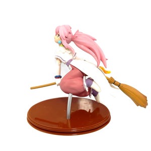 Op de afbeelding: Een beeldje van een personage met roze haar en een wit en roze outfit, rijdend op een bezem. Het beeldje staat in een vliegende pose, met een bruine basis en een lichte achtergrond.