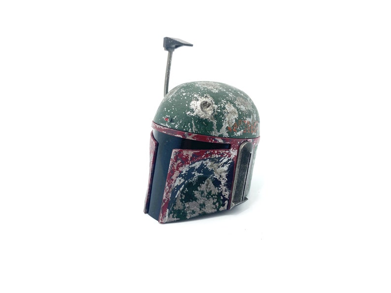May include: A weathered green and red Boba Fett helmet with a black visor and a small antenna on top.