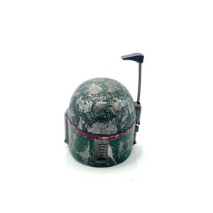 May include: A weathered green and silver Boba Fett helmet with a red stripe and a black antenna. The helmet is a popular collectible for Star Wars fans.