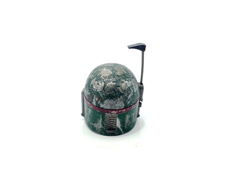 May include: A weathered green and silver Boba Fett helmet with a red stripe and a black antenna. The helmet is a popular collectible for Star Wars fans.
