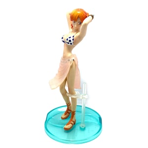 May include: A figurine of a woman with orange hair wearing a pink and white polka dot bikini and a pink sarong. She is standing on a blue and clear plastic base.