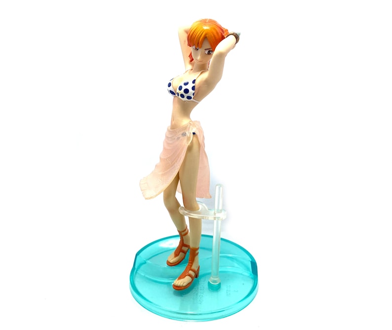 May include: A figurine of a woman with orange hair wearing a pink and white polka dot bikini and a pink sarong. She is standing on a blue and clear plastic base.