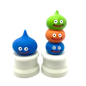 Puede incluir: Tres figuras de slime de colores con caras sonrientes sobre pedestales blancos. Las figuras de slime están apiladas una encima de la otra, con un slime verde en la parte superior, un slime naranja en el medio y un slime azul en la parte inferior.