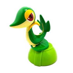 Peut inclure: Jouet en plastique vert et jaune représentant un personnage Pokémon, Grookey, debout sur une base verte.