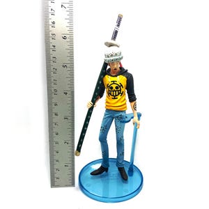 Pu&ograve; includere: Statuetta dettagliata di un personaggio con un top giallo e nero, jeans blu e un cappello bianco con pois neri. La figura tiene in mano un lungo bastone verde scuro e un supporto blu. La figura &egrave; alta circa 15 cm.
