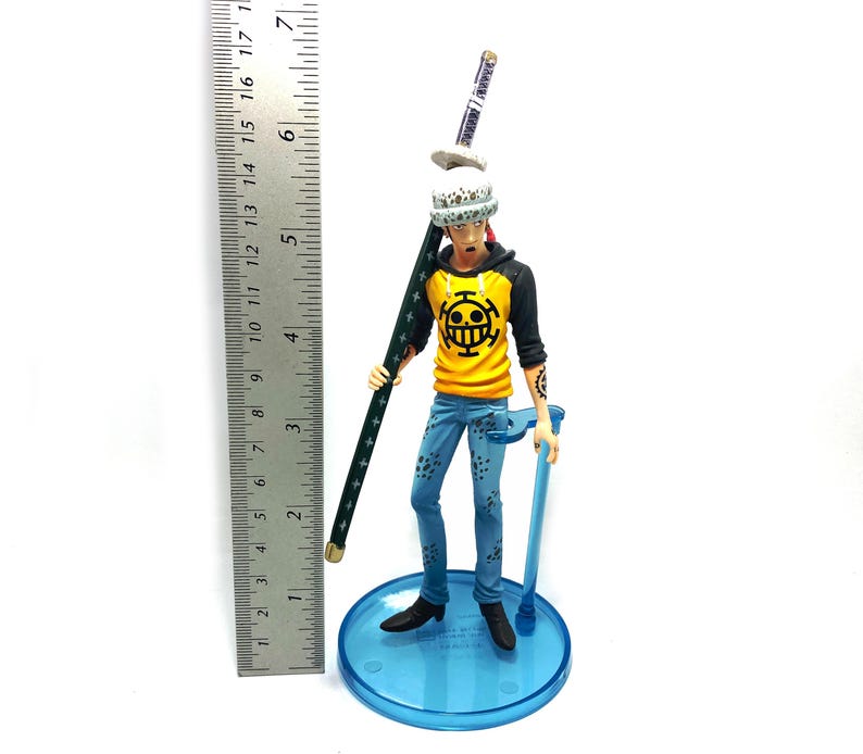 Pu&ograve; includere: Statuetta dettagliata di un personaggio con un top giallo e nero, jeans blu e un cappello bianco con pois neri. La figura tiene in mano un lungo bastone verde scuro e un supporto blu. La figura &egrave; alta circa 15 cm.