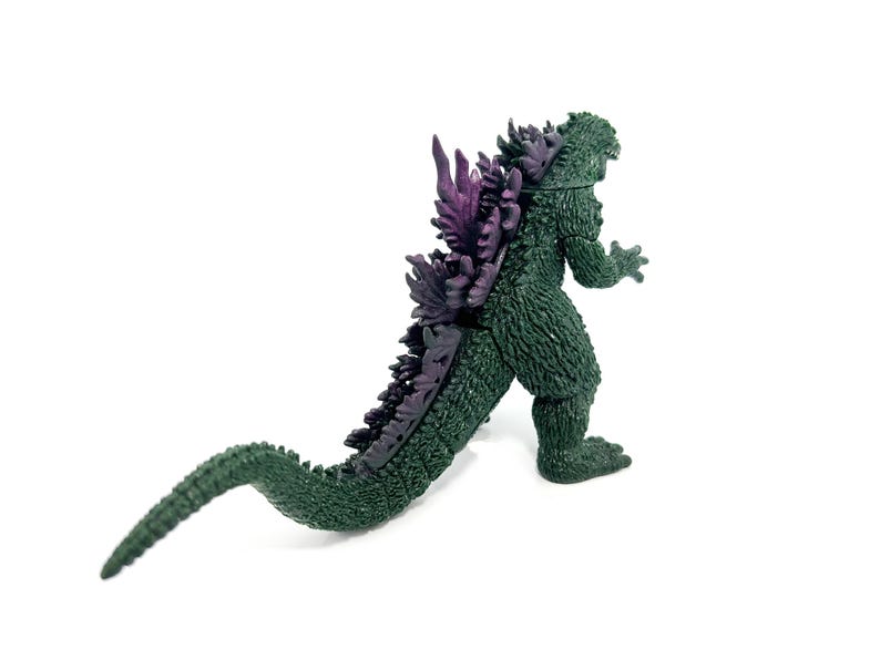 K&ouml;nnte beinhalten: Eine gr&uuml;ne Godzilla-Actionfigur mit lila R&uuml;ckenflossen und einem langen Schwanz. Die Figur ist in einer Laufhaltung dargestellt und zeigt ihre strukturierte Haut und ihr detailliertes Design. Ein Sammlerst&uuml;ck.