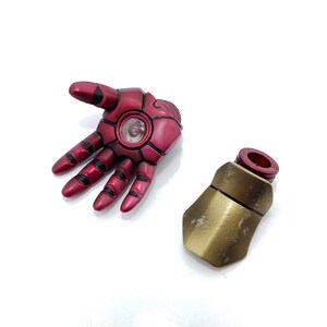 May include: A red and black Iron Man action figure hand with a gold and red forearm piece. The hand has a glowing light in the palm.