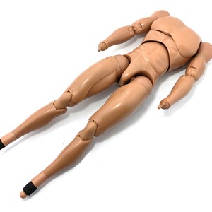 May include: A tan-colored articulated action figure body with a muscular physique. The figure has jointed arms and legs, with black bands around the ankles. The figure is lying on a white surface.