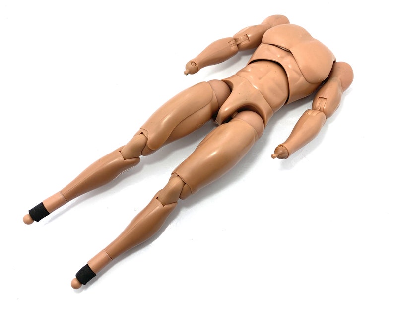 May include: A tan-colored articulated action figure body with a muscular physique. The figure has jointed arms and legs, with black bands around the ankles. The figure is lying on a white surface.