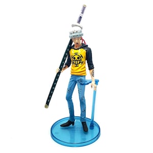 Pu&ograve; includere: Statuetta dettagliata di un personaggio della serie anime One Piece. La figura indossa una felpa con cappuccio gialla e nera con un logo a teschio, jeans blu e un cappello bianco. Tiene una lunga spada verde scuro e si trova su una base blu.