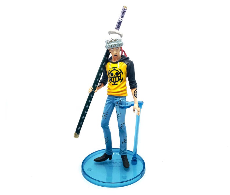 Pu&ograve; includere: Statuetta dettagliata di un personaggio della serie anime One Piece. La figura indossa una felpa con cappuccio gialla e nera con un logo a teschio, jeans blu e un cappello bianco. Tiene una lunga spada verde scuro e si trova su una base blu.