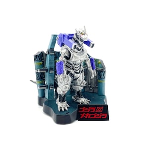 May include: A silver and blue Mechagodzilla action figure stands on a black base with a blue and teal backdrop. The figure has a detailed design and the Japanese text "Godzilla X Mechagodzilla" is on a red and black label.